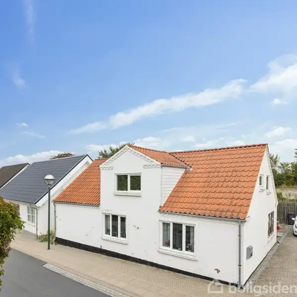 Hvidt hus med rødt tegltag står stille på en villavej omgivet af andre boliger og biler, under en klar blå himmel. En smal flisebelagt sti løber langs huset.