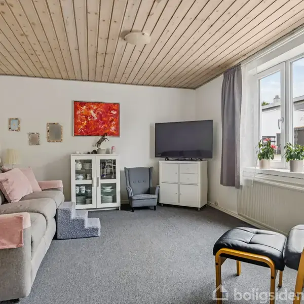 Gråt sofaarrangement med lyserøde puder og tæpper står i stue med træloft. En TV-skærm står på kommode ved siden af en glasvitrine. Et stort vindue giver naturligt lys.