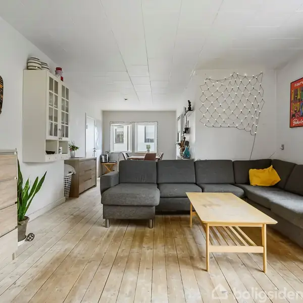 Grå hjørnesofa med puder står i en stue med træplankegulv og lyse vægge. Et sofabord foran sofaen, malerier og planter dekorerer rummet. Stuen har også indbygget hylde og spisestole i baggrunden.