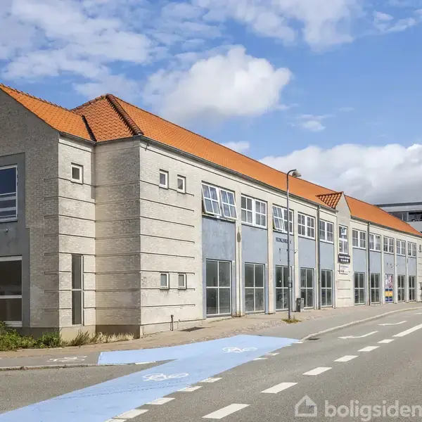 En lysgrå bygning med rødt tegltag, store vinduer, og skilt ved indgangen. Den står ved en vejkryds med en cykelsti i nærheden. Skyer og blå himmel ses i baggrunden.