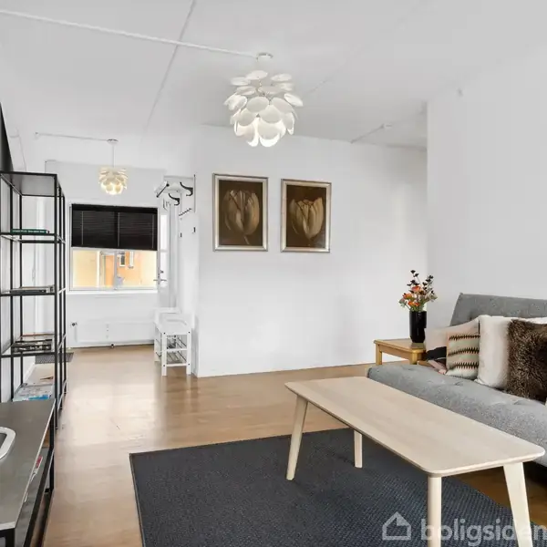 Stue med grå sofa og sofabord, dekoreret med puder på en trælaminatgulv. Væggene er hvide, to billeder hænger, og der er en reol ved siden af et tv.