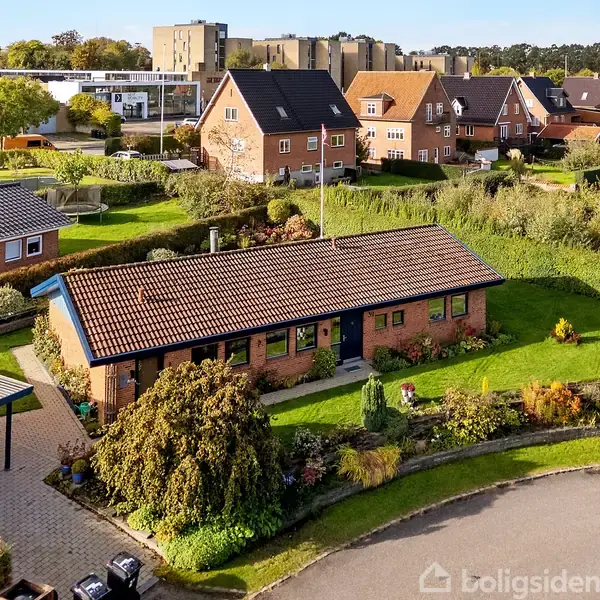 Rødstenshus med blåt træværk ligger i en velplejet have med blomsterbede, omgivet af andre villaer og et grønt område i et villakvarter.