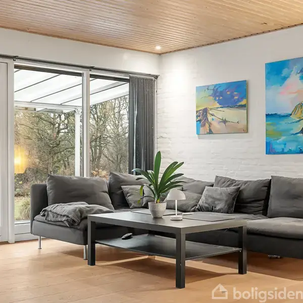 Grå sofa står i stue med lyse trægulve, foran et sort sofabord med en plante. Store vinduer viser en udendørs have, og to farverige malerier hænger på væggen.