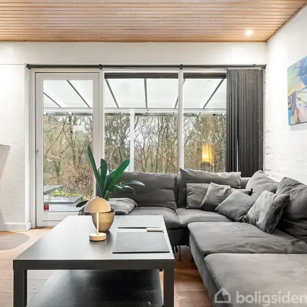 En grå hjørnesofa står i en stue ved et stort vindue, med udsigt til træer. På bordet er en lampe og bøger. Et blødt lys fylder rummet, og der hænger malerier på væggen.