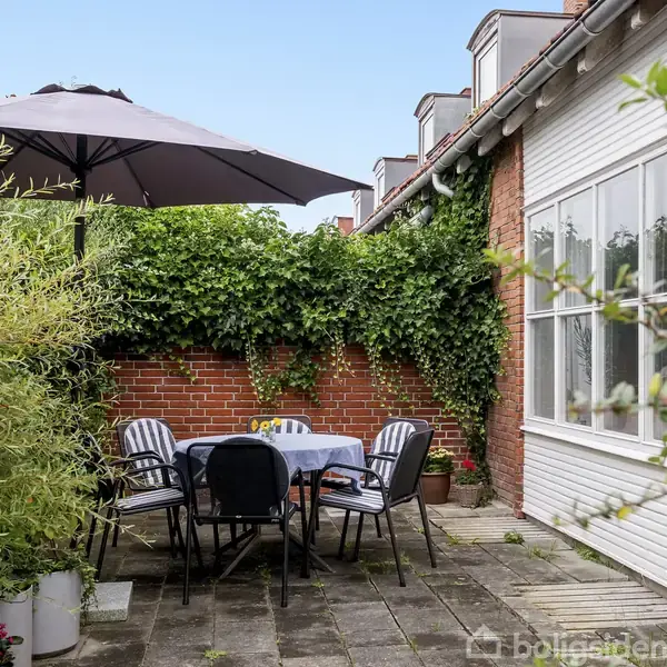 Et udendørs terrasseområde med et bord og fem stole under en parasol. Omgivet af murstensbygninger og grønne planter, skaber en hyggelig atmosfære.