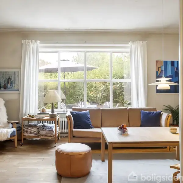 Stue med lysebrun sofa og blå puder, placeret foran et stort vindue med udsigt til en have. Et sofabord foran sofaen omgivet af lænestole og en lampe.