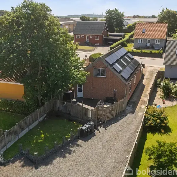 Hus med solpaneler på taget omgivet af have og hegn. Beliggende på en grusvej i et villakvarter med andre huse og haver. Træer og græsplæne udgør baggrundslandskabet.