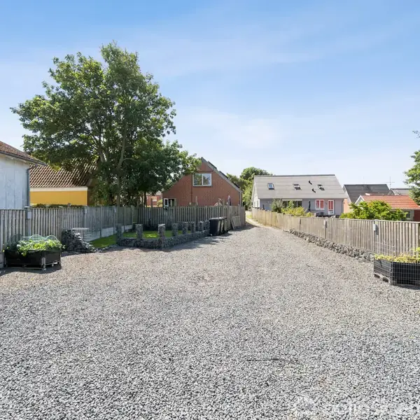 En grusvej fører gennem rækkehuse, kantet af træhegn og containere med planter. Omkranset af grønne træer og lyseblå himmel.