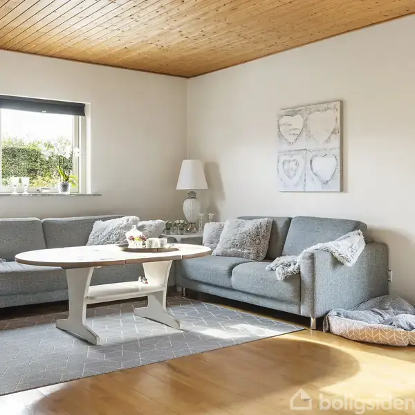 En grå hjørnesofa står stille i en stue med trægulv. Sofaen har puder, et tæppe ved siden af, et lavt sofabord foran og en lampe ved siden.
