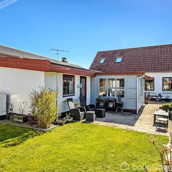 Et hus står stille i en velholdt have med grøn græsplæne og flisebelagt terrasse. Omkring huset er der en klar blå himmel og solskin.