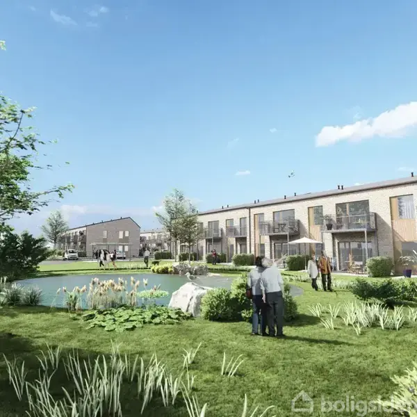 Boligbebyggelse langs en sø i et grønt område; to personer står nær vandet, og flere moderne rækkehuse er synlige i baggrunden under en klar himmel.