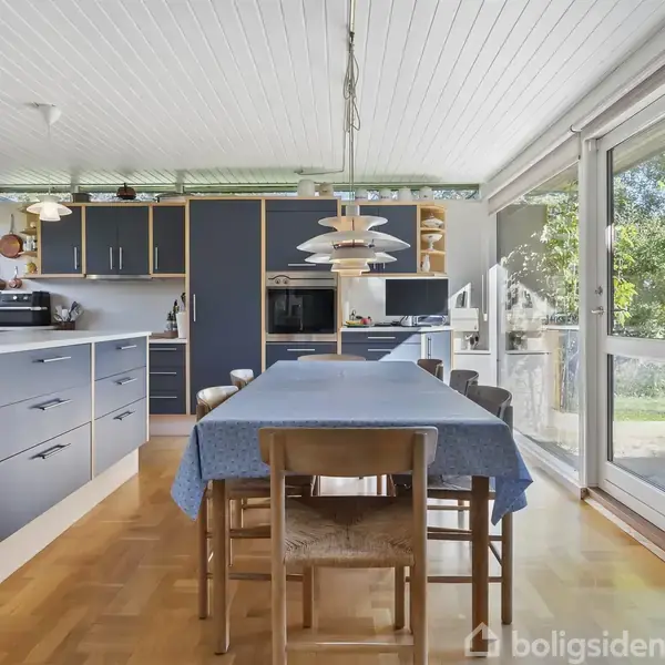 Et spisebord med blå dug står i en moderne køkken-alrum med blå skabslåger. Rummet er lyst med store vinduer, der vender ud mod en grøn have med havemøbler.