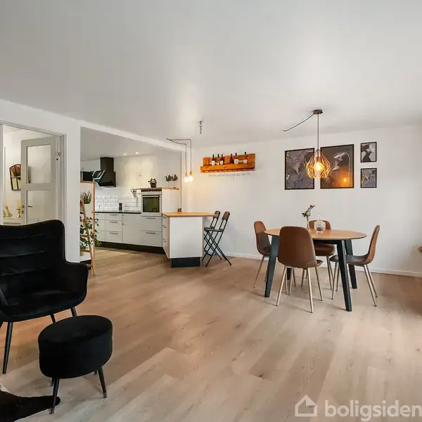 Stuen har et moderne design med et sort lænestolssæt og en rund spisebordgruppe. I baggrunden ses et åbent køkken og hvidmalede vægge med enkelte plakater.