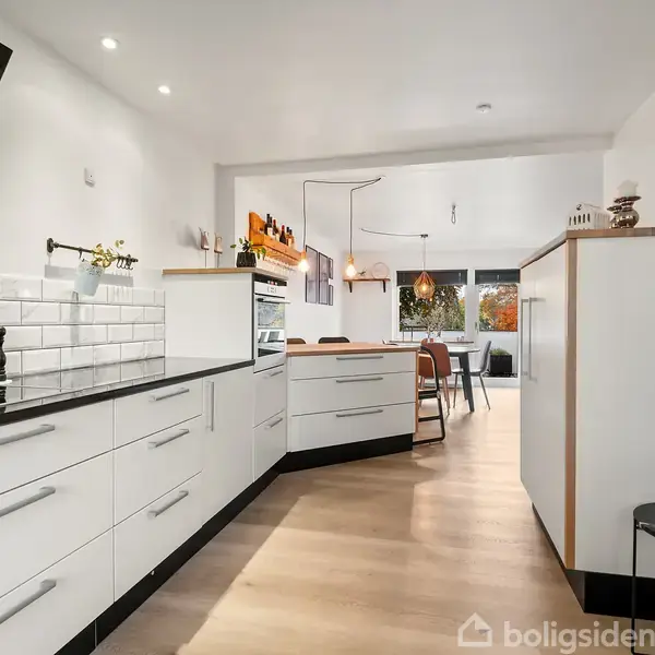 Moderne køkken med hvide skabe og sorte bordplader. Hvide fliser på væggen ved komfuret. I baggrunden ses spiseområde med vinduer, der giver naturligt lys.