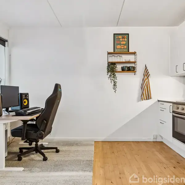 Kontorstol ved skrivebord med computer set ved et stort vindue. Til højre ses et køkken med hvide skabe og ovn. En plante hænger fra en lille hylde på væggen.