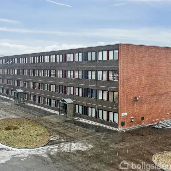 En lang, tre-etagers murstensbygning står stille omgivet af en åben gårdsplads med græsarealer og stier. Skyerne i himmelen antyder overskyet vejr. Ingen tekst ses i billedet.