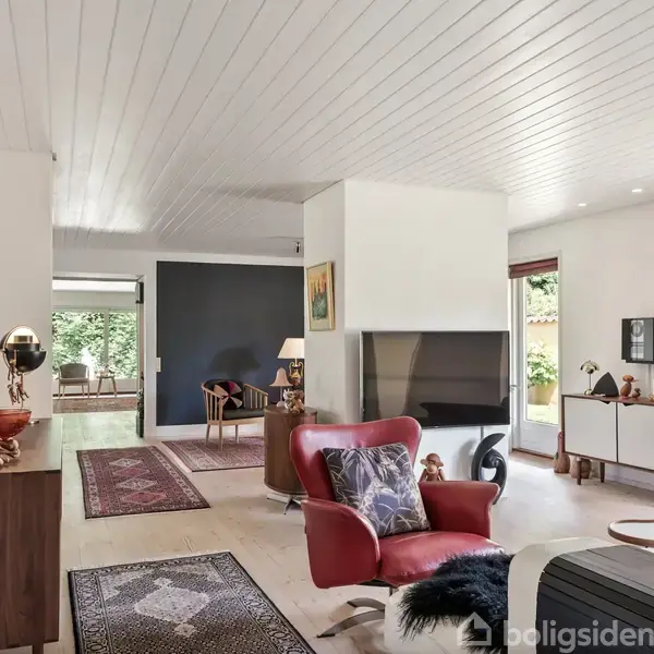 En rød lænestol står i en lys stue med hvidt loft og parketgulv. Rummet har store vinduer, tæpper og moderne møbler. En pejs står centralt i rummet.