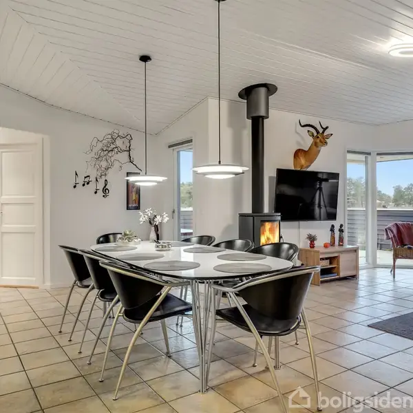 Spisebord med sorte stole er placeret i et lyst rum med klinkegulv. Der er en pejs ved siden af en fladskærms-TV, dekorativ hjortehovedmontering og store vinduer mod en terrasse.