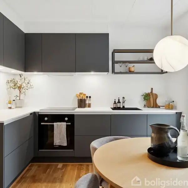 Moderne køkken med sorte skabe og hvide bordplader, hvor en rund spisebord med sorte og grå detaljer står. Et ovn og komfur indbygget, planter og flasker pynter rummet.