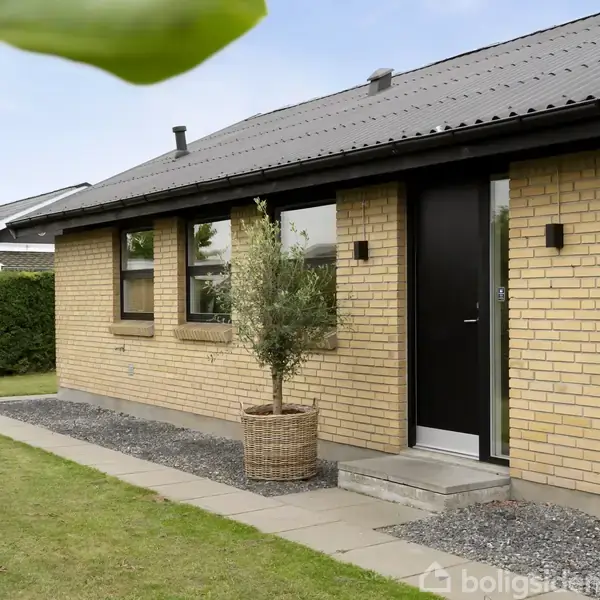 Et gulstenshus med sorte vinduesrammer og en sort dør står stille. Foran indgangspartiet er der en plante i en stor kurv. Huset omringes af en velplejet græsplæne og hække.