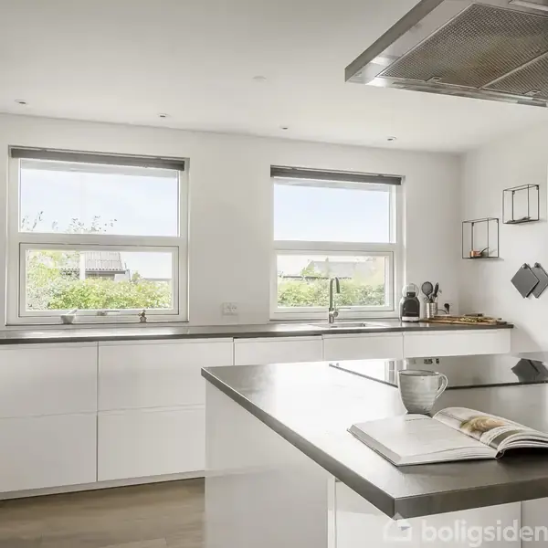 Et moderne køkken med en hvid bordplade, vask og vinduer. En åben bog og en kop står på køkkenøen. Lyse omgivelser fylder rummet.