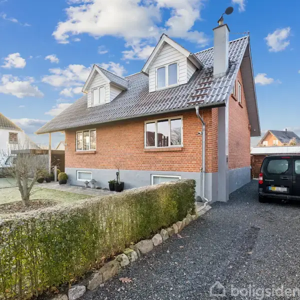 Et rødt murstenshus med en grusbelagt indkørsel og sort bil parkeret ved siden af. Huset har gavle med vinduer og en frodig have med hæk i forgrunden.