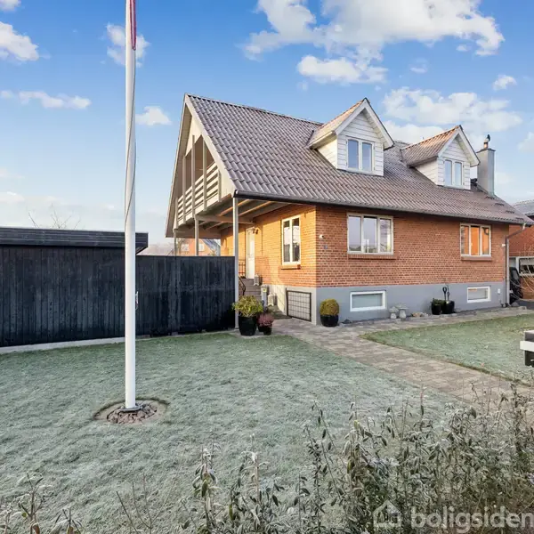 Et rødt murstenshus med skråt tag står i en have med frost på græsset. Til venstre ses en flagstang og et sort skur.