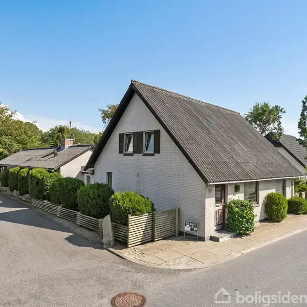 Huset, en gammel villa, står på et hjørne i et roligt nabolag. Det har en A-formet tag og hvide mursten, omkranset af velplejede hække og træer langs asfalterede veje.