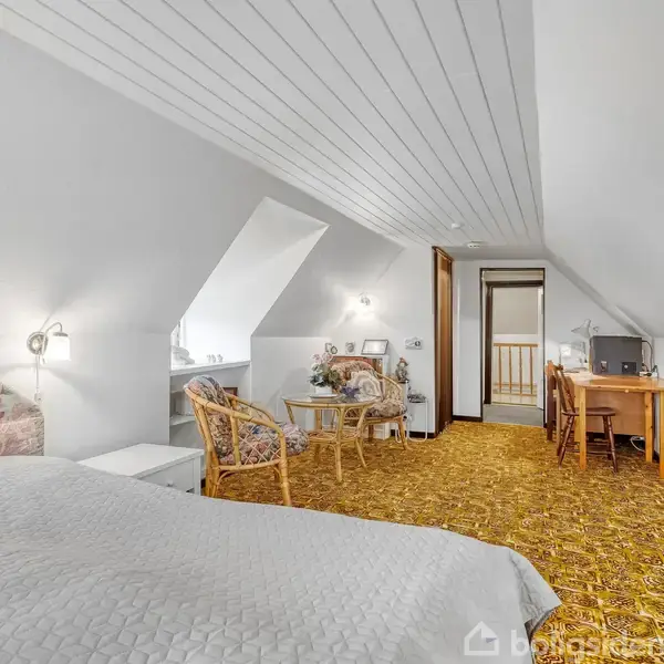 Soveværelse med skråtag, hvor en seng er placeret til venstre. To stole og et bord står ved et vindue. Et skrivebord med stol er nær døren.