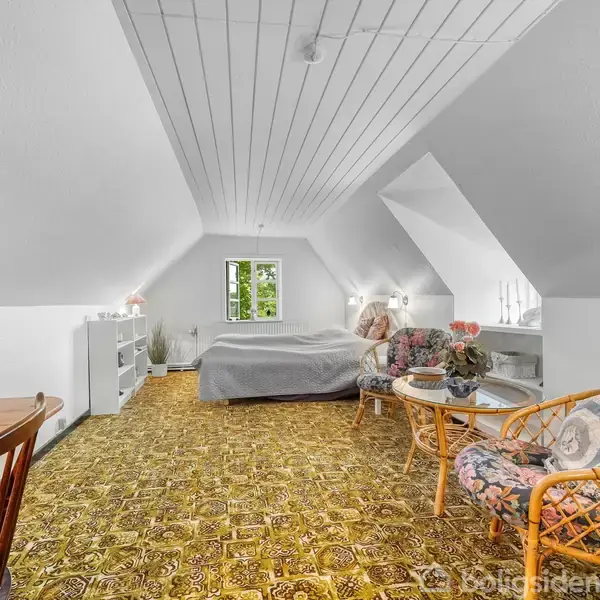 Soveværelse med skrå loftsvægge indeholder en seng mod væggen, to kurvestole og et lille bord med blomster. Gulvtæppet er mønstret. Et bord med en stol er til venstre.