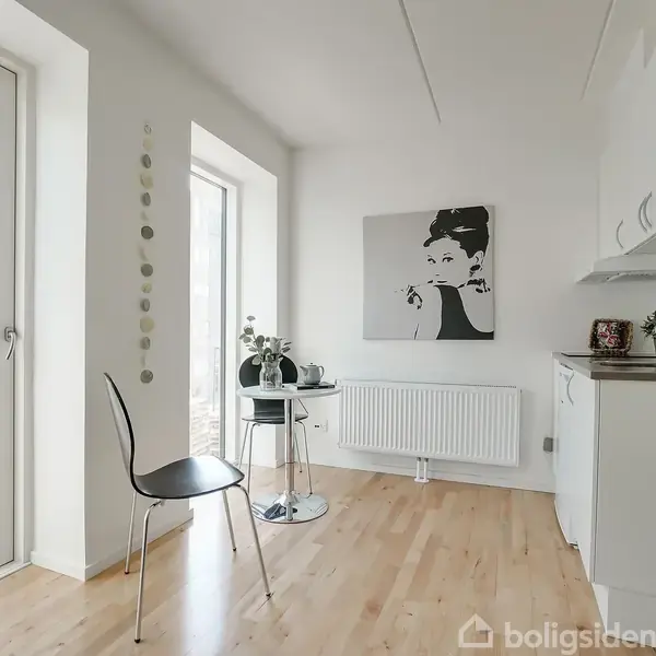 En moderne køkken-stue med lyst trægulv indeholder en sort stol og et rundt bord med planter. Store vinduer giver udsigt til en boligblok. En væg har et sort-hvidt portræt.