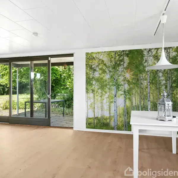 Et lyst rum indeholder et hvidt bord med lanterne; store glasdøre åbner op til en have, og en væg er dekoreret med et skov-inspireret tapet.