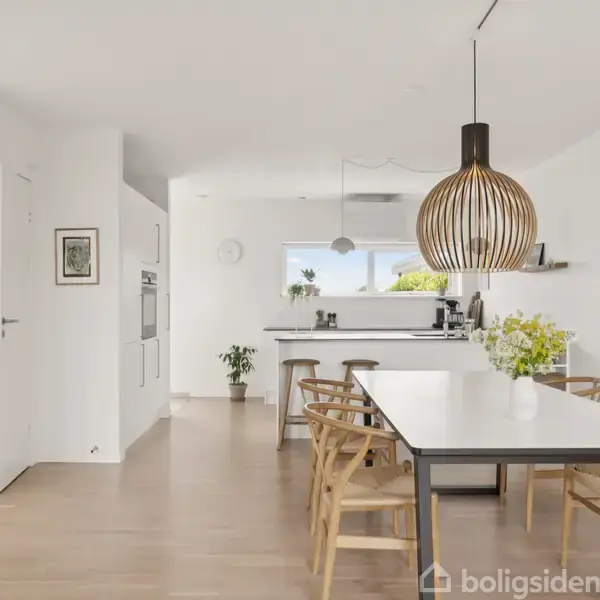 Et moderne køkken-alrum med et stort spisebord omgivet af stole. En minimalistisk lampe hænger over bordet. Baggrunden viser et hvidt køkken med planter og lyse træelementer.