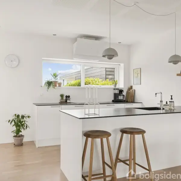 Moderne køkken med en central ø, to høje taburetter og en sort bordplade. Rummets hvide vægge og store vindue skaber en lys atmosfære, suppleret af hængende lamper og planter.