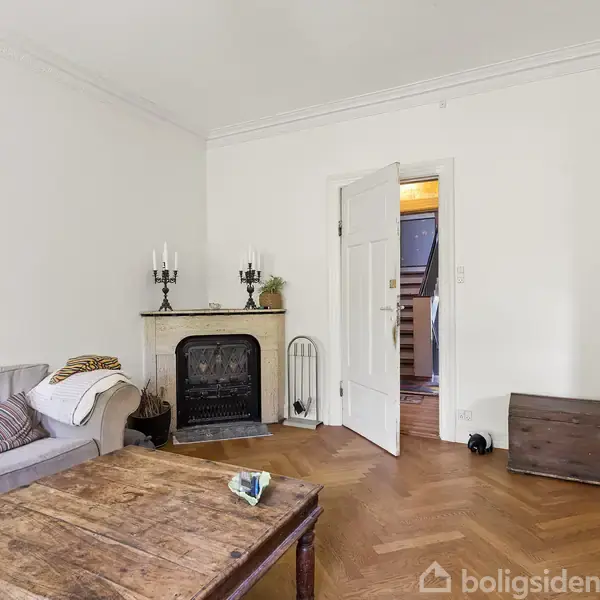 En grå sofa står i en stue med en pejs, hvor to lysestager står ovenpå. Et træbord står foran sofaen. En åben dør fører til en gang.