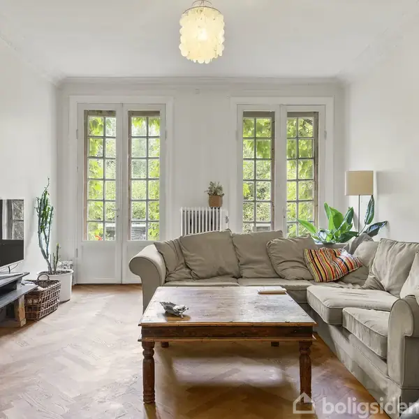 En grå sofa står i en lys stue med store vinduer, som slipper grønt løv udenfor ind. Et træbord er foran sofaen. Fjernsynet står på en lav reol til venstre.