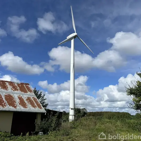 Vindmølle drejer langsomt ved siden af en gammel bygning med rustent tag i et landligt landskab. Baggrunden viser blå himmel med spredte skyer.