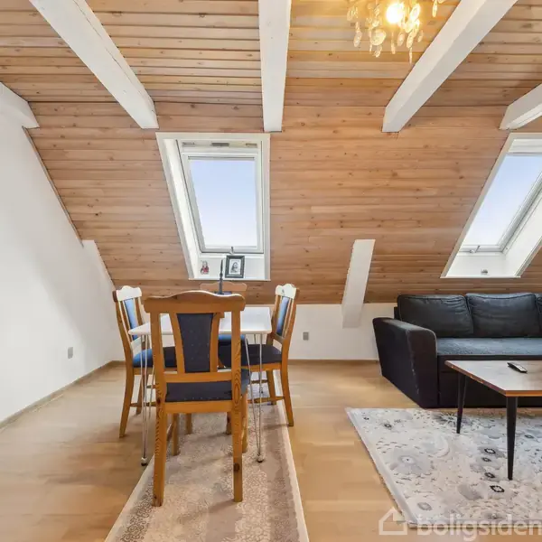 En spisebord oplevelse med fire stole står i et rum med skråtloft og loftsvinduer. En sofa og tv-bord ses i stuen, dekoreret med kunstværker på væggen.