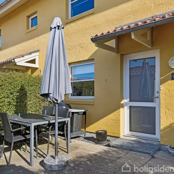 Et gult rækkehus har en terrasse med et bord, stole og en parasol. Dørskilt viser nummer "134". Omkringliggende hæk giver en privat haveatmosfære.