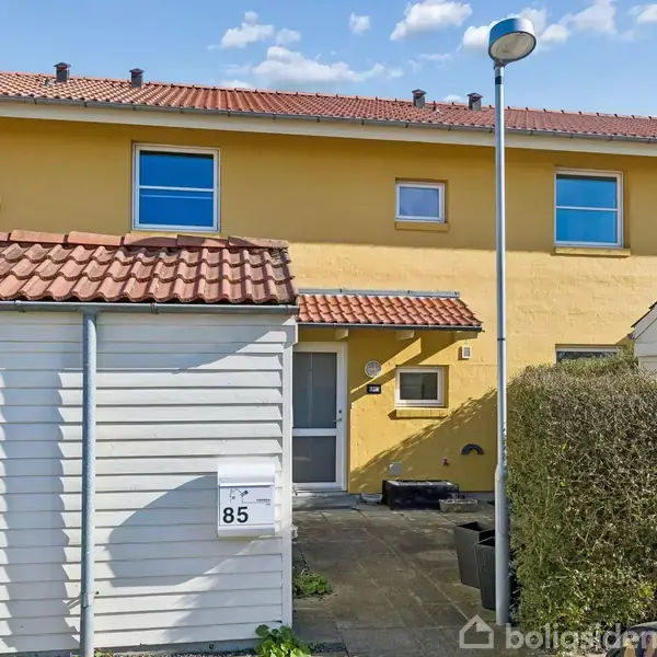 Hus med gule mure og to etager, set forfra. Hoveddøren under et lille halvtag. Hvide skure med nummer 85 og flisebelagt forhave omkranset af buske.