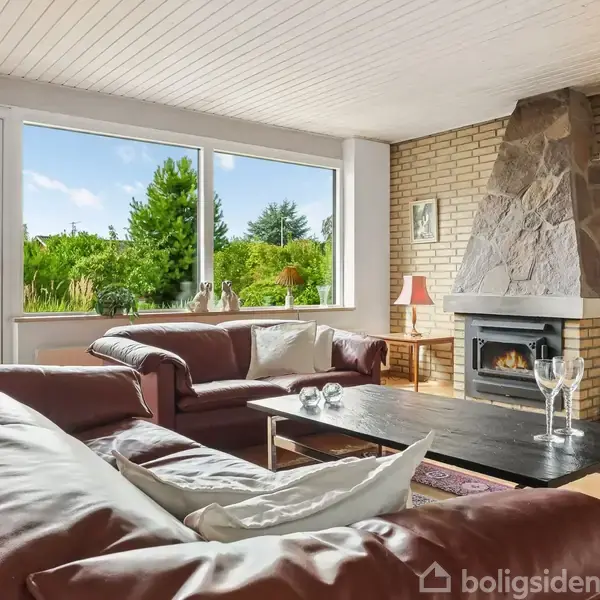Brun lædersofa står i en stue med store vinduer. En pejs er tændt ved en murstensvæg. Lyse trægulve, et bord med glas, og udsigt til grøn have.