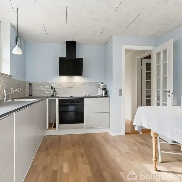 Køkken med moderne grå skabe, komfur og emhætte. Bordplade med vask ved vindue. Spisebord med dug står til højre. Trægulv og hvide vægge kompletterer det lyse rum.
