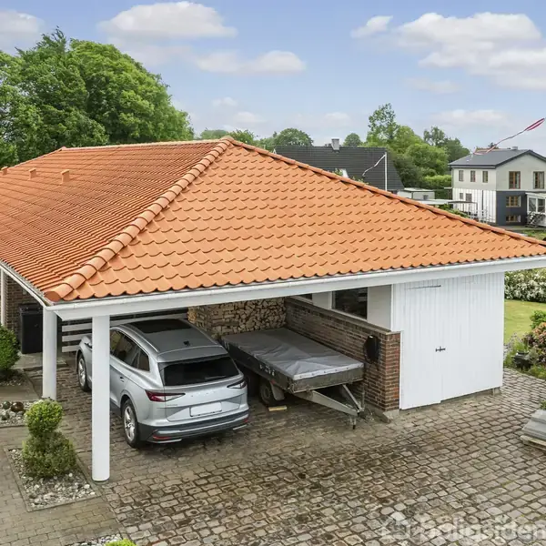Carport med rød teglstenstag, der beskytter en hvid bil parkeret ved siden af en trailer. Omgivet af en have og andre huse i et villakvarter.