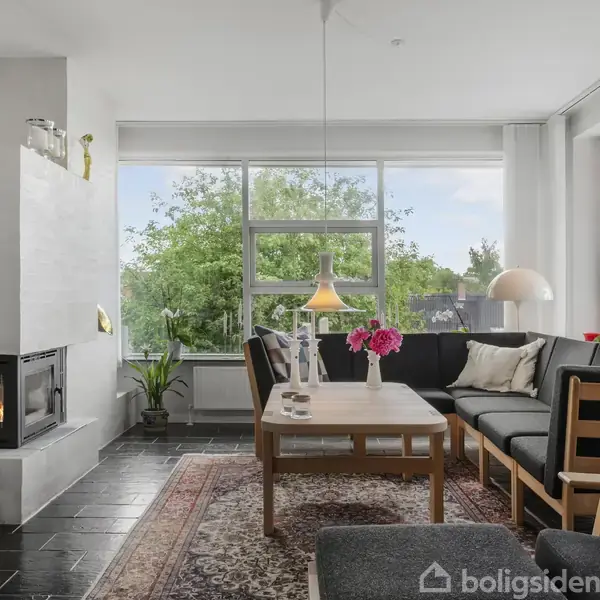 Brændeovn ved siden af et sofabord med blomster og omgivet af en hjørnesofa. Vinduer viser grønne træer udenfor, og rummet har stilfulde lamper og tæpper.