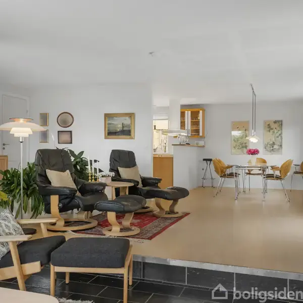 Stue med sort lænestol og fodskammel foran et sofabord. I baggrunden ses spisebord med stole, lamper og planter. Billeder hænger på væggen i rummet med lyse vægge.