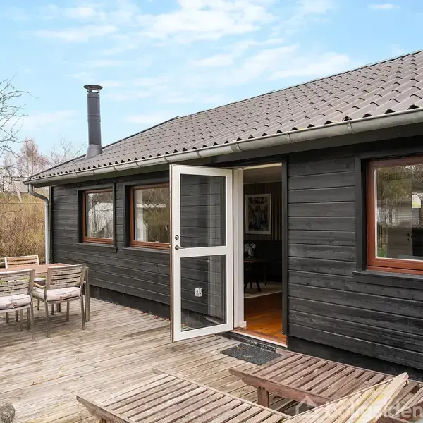 Sort træhus med åbent vindue og dør, placeret på en træterrasse. Terrassebord med stole og parasol i baggrunden. Flere træer og buske i baggrunden.