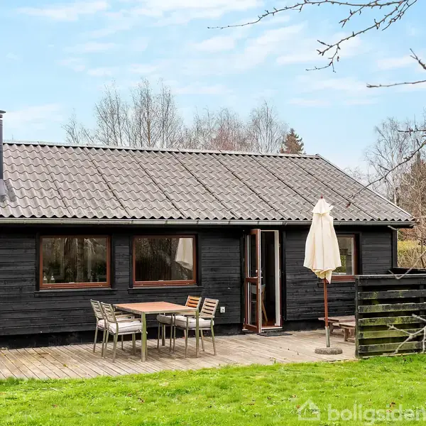 Sort træhus står stille på græsplæne omgivet af træer. På terrassen foran huset er der et bord med stole og en paraply.
