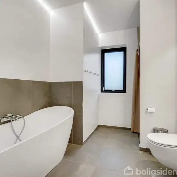 Et moderne badeværelse med et fritstående badekar til venstre, en toiletpapirholder ved siden af toilettet til højre, og en lille sort vase med blomster på en hylde i baggrunden.