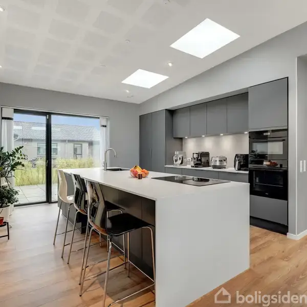 Køkken med stor hvid køkkenø; flere barstole står omkring. Gråt indbygget skab med indbygningsovn og apparater. Store vinduer leder ud til en terrasse med planter.