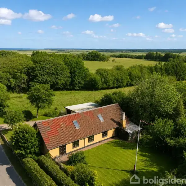 En enkeltstående gul bygning med rødt tag ligger i en frodig, grøn landskabelig omgivelser ved siden af en vej. Træer og marker strækker sig i baggrunden under en blå himmel.